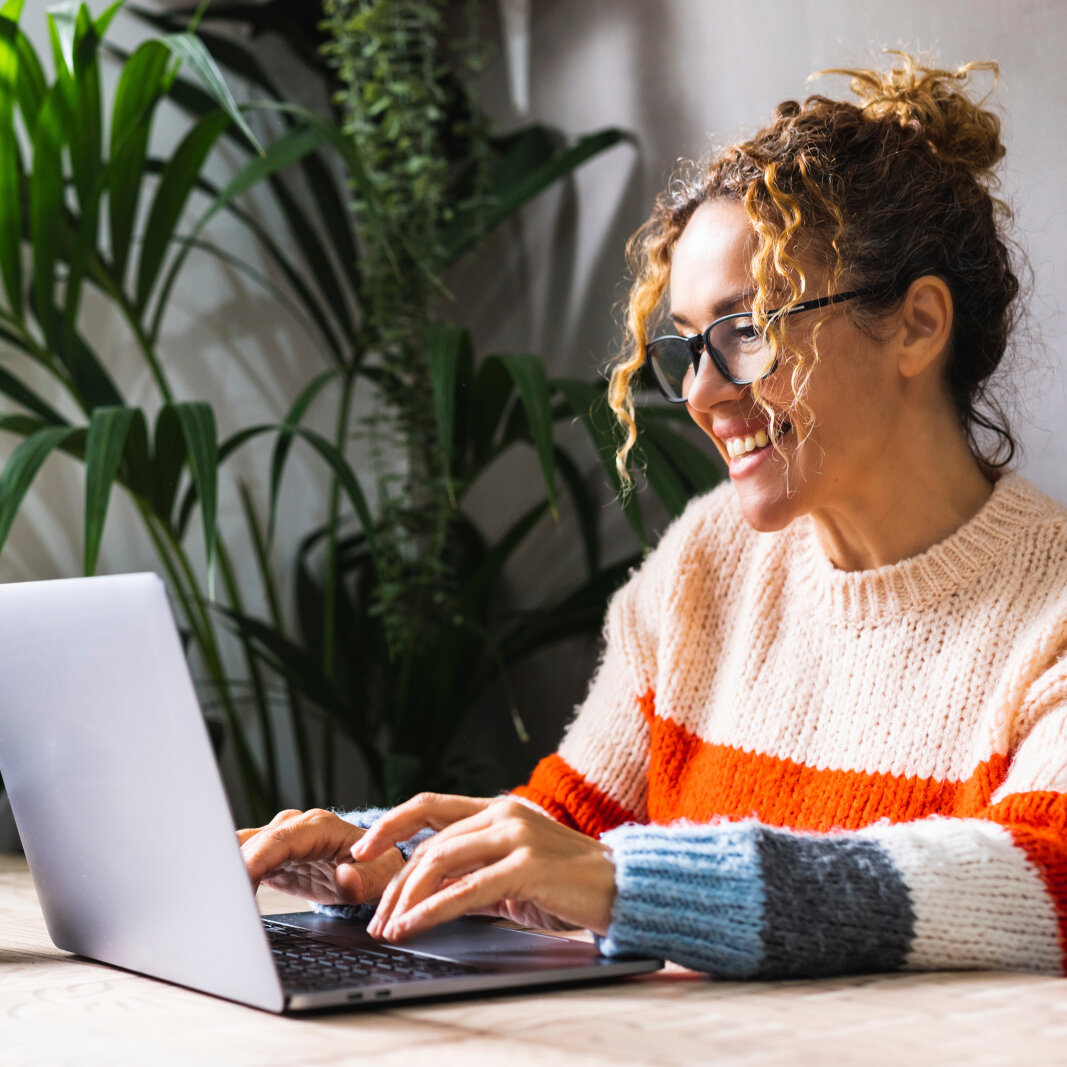 Happy woman writing on her laptop in a room with plants Happy woman writing on her laptop in a room with plants